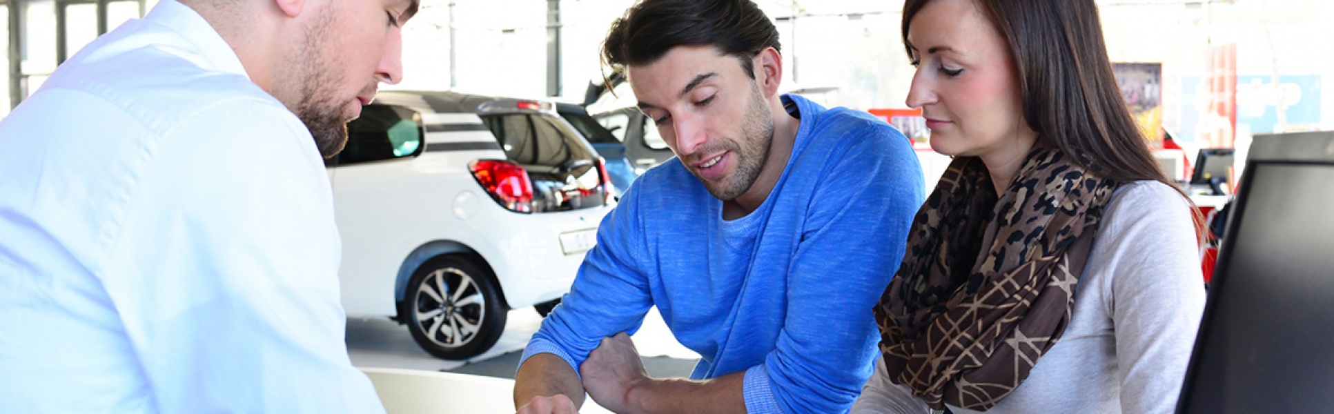 Mann und Frau im Gespräch am Schreibtisch mit Kundenberater, im Hintergrund Ausstellungsfahrzeuge